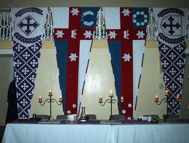 Standards for the Kingdom of Lochac, and the Baron and Baroness of St Florian de la Riviere on display indoors behind a high table at a feast
